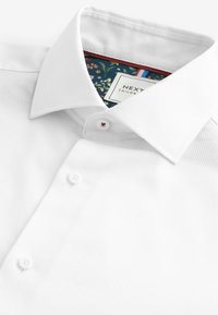 Camisa de vestir blanca texturizada con cuello amplio, que presenta un interior con estampado floral y dos cierres de botón en el frente.