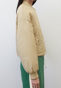 Giacca bomber beige con una texture liscia, polsini a coste e una tasca laterale con zip, caratterizzata da una vestibilità comoda e un orlo arrotondato.