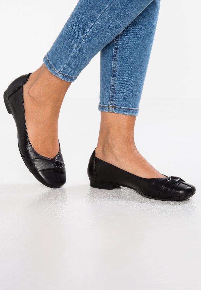 Ballerines en cuir noir avec un détail noué sur le bout, talon plat, et une finition lisse et brillante. Portées avec un jean bleu ajusté.