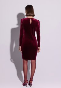 Burgundowa aksamitna sukienka z długimi rękawami, z klamrą na plecach, dopasowanym fasonem, zestawiona z pasującymi butami na obcasie i przejrzystymi pończochami.