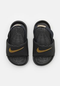 Černé skládací sandály s širokým páskem, který má zlaté logo Nike. Texturovaná černá gumová podrážka má kruhové vzory pro lepší přilnavost.