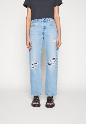 Personne portant un jean droit déchiré bleu clair, des sandales noires et une chemise sombre à manches courtes, debout devant un fond uni.
