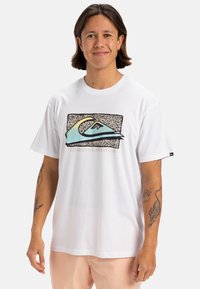 Camiseta de algodón blanca con un gráfico de olas multicolores sobre un fondo de patrón en blanco y negro, con el texto "Quiksilver" debajo.