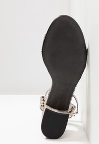 Sandale à bout ouvert avec une tige beige texturée et une semelle en caoutchouc noire. Comprend des sangles réglables et un logo de marque embossé sur la semelle.