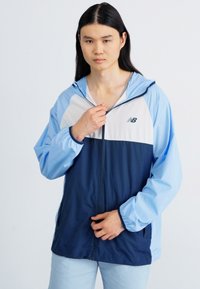 Lahka jakna na zadrgo v svetlo modri in mornarsko modri barvi, z barvnim blokiranjem, nastavljivo kapuco in logotipom New Balance. Gladka tekstura.