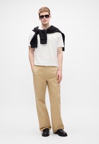 Camiseta branca de manga curta, calças largas bege, suéter preto amarrado ao redor do pescoço, óculos de sol pretos e sapatos pretos em um fundo plain.