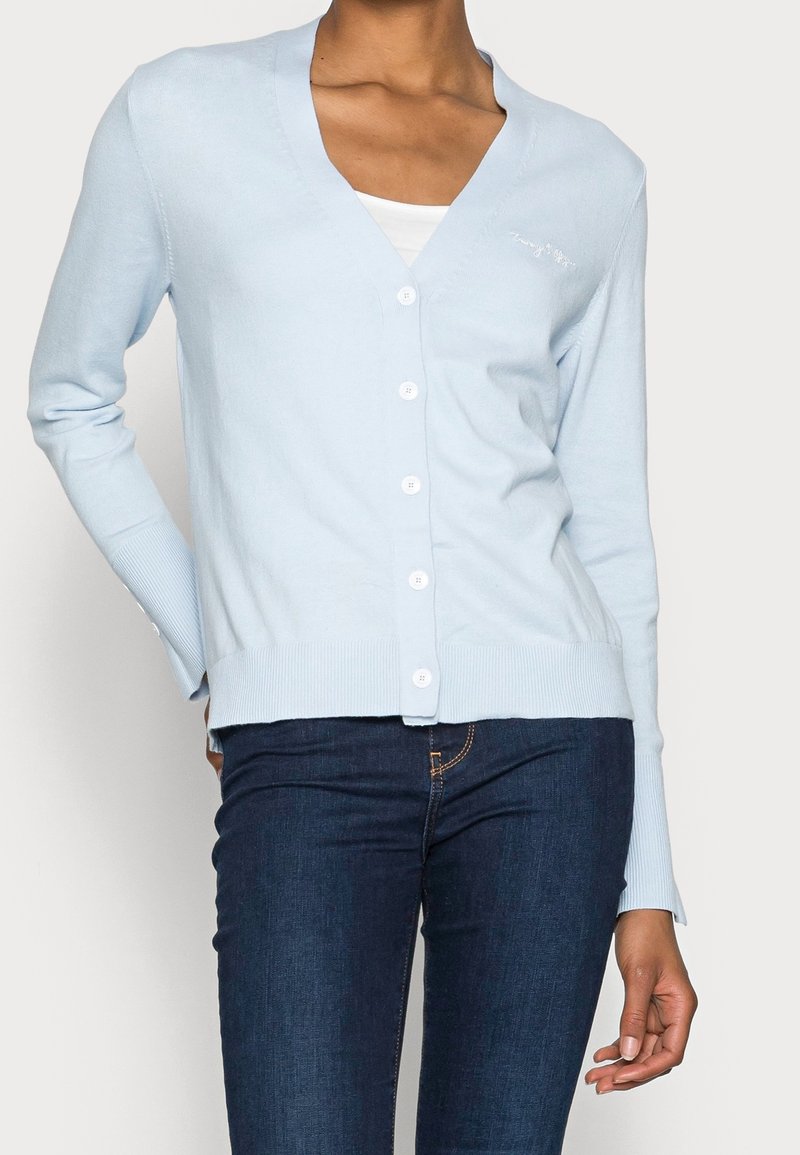 Mujer con un cárdigan abotonado azul claro sobre una blusa blanca y jeans azul oscuro, contra un fondo liso.