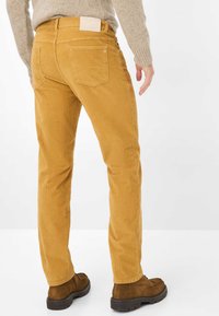 Homme portant un pantalon en velours côtelé couleur moutarde, un pull beige et des bottines en daim marron, debout devant un fond blanc uni.