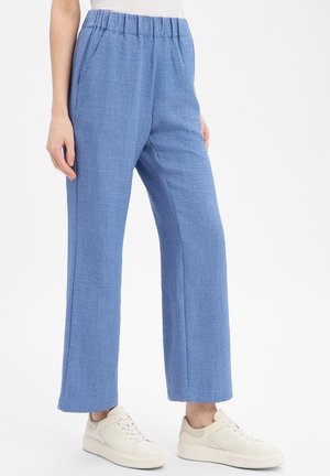 Frau trägt hellblaue, strukturierte, weit geschnittene Hosen mit elastischem Bund und weißen Turnschuhen, steht vor einem weißen Hintergrund.