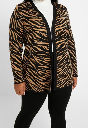 Woman wearing a black and tan tiger-striped cardigan over a white ribbed turtleneck, paired with black leggings.