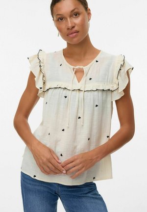 Blouse crème clair avec des manches à volants, motifs en forme de cœur noirs, décolleté en trou de serrure et détail à cordon de serrage. Tissu texturé.