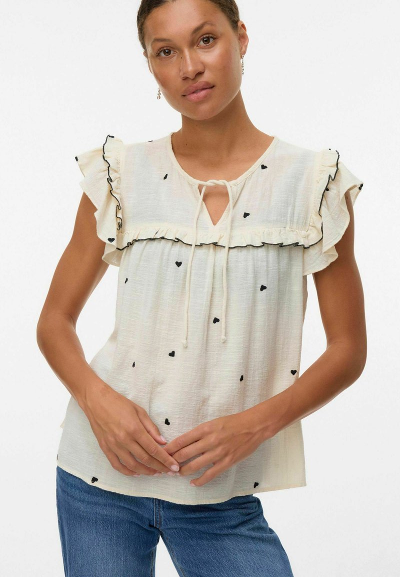 Blouse crème clair avec des manches à volants, motifs en forme de cœur noirs, décolleté en trou de serrure et détail à cordon de serrage. Tissu texturé.
