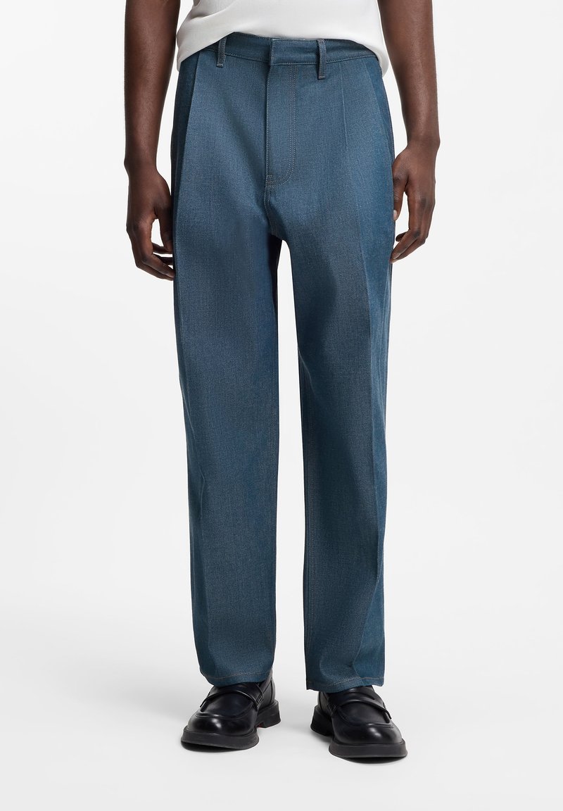 Άνδρας που φοράει μπλε παντελόνι με ίσια γραμμή και μαύρα δερμάτινα παπούτσια, στέκεται μπροστά σε άσπρο φόντο, με τα χέρια χαλαρά δίπλα στο σώμα.