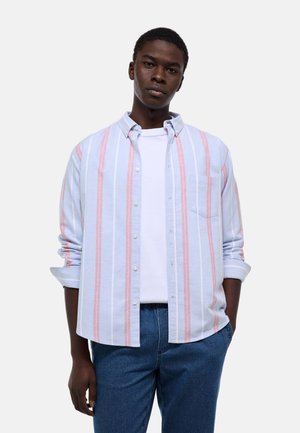 Jeune homme portant une chemise à rayures verticales bleu clair et rose sur un t-shirt blanc et un jean bleu, debout avec une main dans la poche.