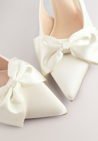 Paire de chaussures pointues en satin blanc avec de grands nœuds décoratifs en satin à l'avant, montrées en gros plan sur une surface claire.