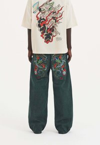 Krémová košile s barevným potiskem lebky a plamenů na zadní straně, spárovaná s tmavě zelenými širokými džínami se složitým vyšíváním draka.