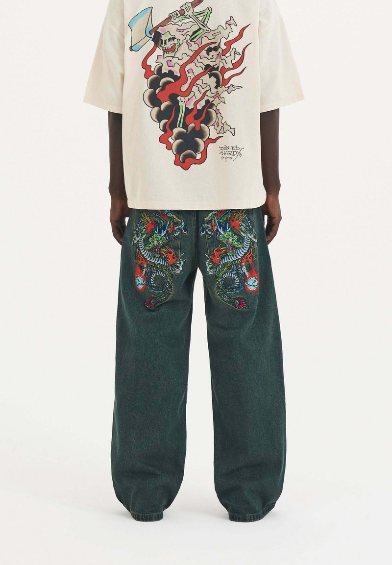 Krémová košile s barevným potiskem lebky a plamenů na zadní straně, spárovaná s tmavě zelenými širokými džínami se složitým vyšíváním draka.
