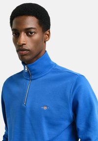Pullover blu con zip e colletto strutturato in morbido materiale felpato, caratterizzato da un piccolo logo sul lato sinistro del petto. Texture del tessuto liscia.