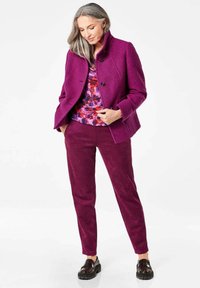 Paarse wolmix jas met een staande kraag, dubbele knopen, gecombineerd met een kleurrijke bloemenblouse en bijpassende paarse broeken.