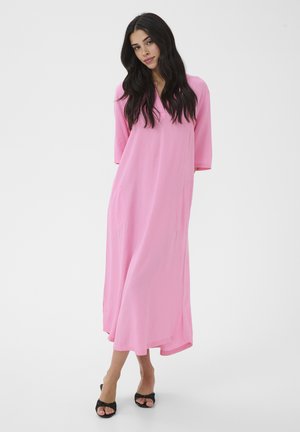 Femme aux longs cheveux noirs portant une robe ample rose vif jusqu'aux chevilles et des talons ouverts noirs, posant devant un fond blanc uni.