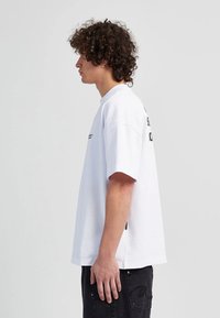 Weißes Baumwoll-T-Shirt mit kurzen Ärmeln, ausgestattet mit einem Rundhalsausschnitt und schwarzem Drucktext auf der Rückseite, kombiniert mit schwarzen Jeans.