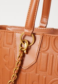 Bolso de mano texturizado en marrón con un diseño estructurado, herrajes dorados y un patrón de logo embossado. Incluye un detalle de cadena en la correa.