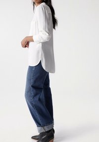 Chemise blanche à manches longues avec une poche, associée à un jean bleu taille haute et des bottines noires à talon en bois, présentée de profil.