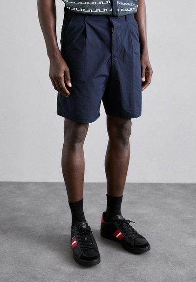 Shorts bleu marine en tissu léger, dotés de plis et d'une fermeture éclair, associés à des baskets noires et des chaussettes noires côtelées.