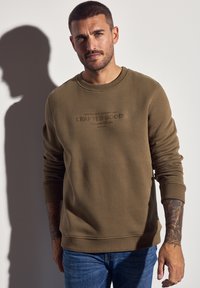 Mand med kort hår og tatoveringer iført en brun sweatshirt med teksten "Crafted Goods," parret med blå jeans, står mod en hvid væg.
