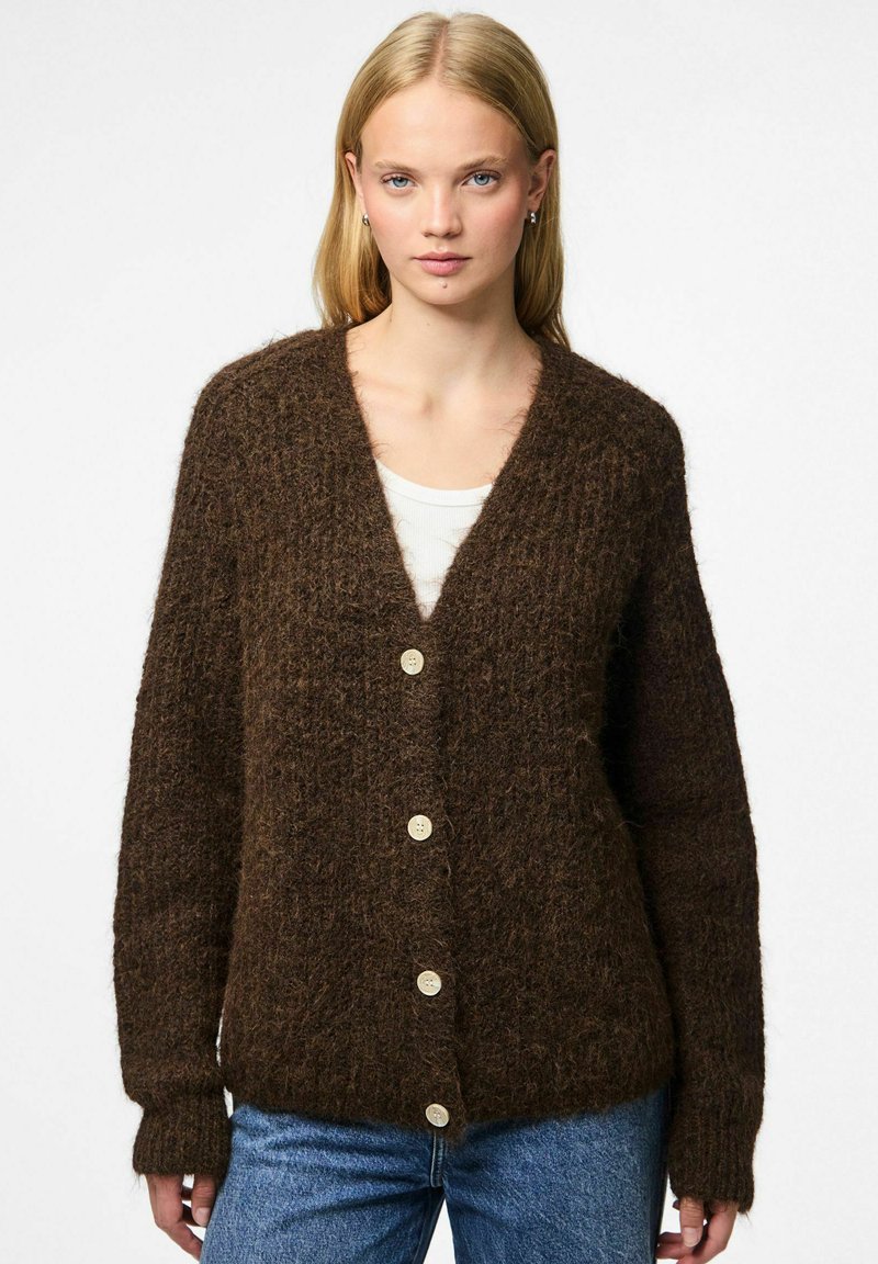 Noor naine, kes kannab pruuni siledat nööbiga kardigani valge pluusi ja siniste teksade peal, seisab lihtsa heleda tausta ees.