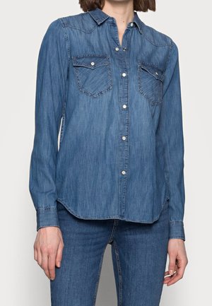 Vrouw draagt een lange mouw blauwe denim blouse met klepzakken op de borst en bijpassende blauwe denim jeans, staand tegen een effen achtergrond.
