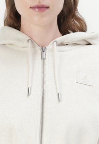 Licht beige rits-hoodie van zachte stof. Bevat een zilveren rits, koordjes en een klein logo patch op de linkerborst.