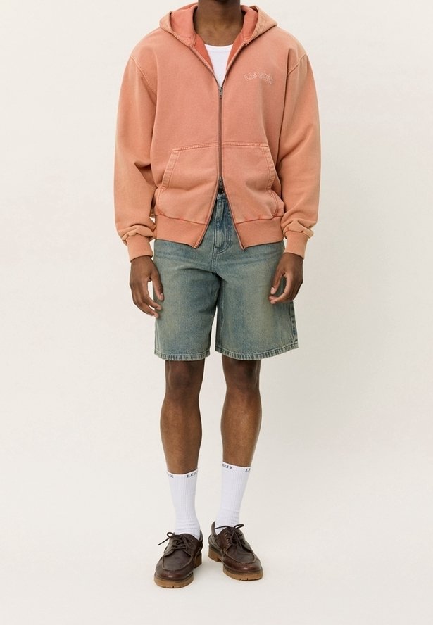 Homme portant un sweat à capuche pêche zippé, un short en jean bleu clair, des chaussettes blanches montantes et des chaussures en cuir marron, debout devant un fond uni.
