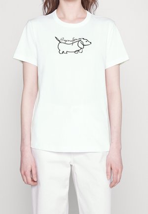 Kobieta nosząca białą koszulkę i spodnie z czarnym konturem jamnika i kursywnym napisem na piersi.