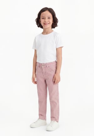 Enfant aux cheveux bouclés foncés portant un t-shirt blanc, un pantalon rose et des baskets blanches, debout et souriant sur un fond blanc uni.