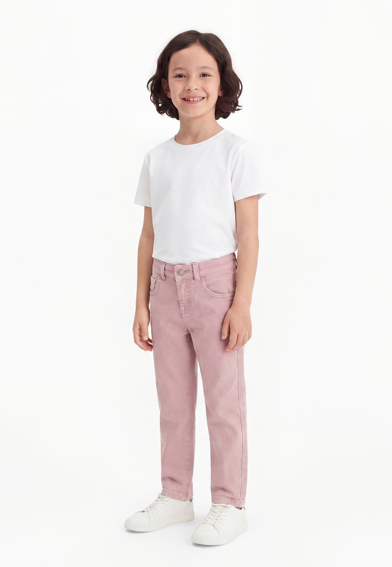 Enfant aux cheveux bouclés foncés portant un t-shirt blanc, un pantalon rose et des baskets blanches, debout et souriant sur un fond blanc uni.