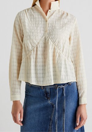 Femme portant un chemisier crème texturé à manches longues avec un col montant et un devant boutonné, associé à une jupe en denim bleu nouée.
