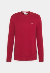 Maglione rosso lavorato a maglia con maniche lunghe, collo rotondo, polsini e orlo a costine. Presenta un piccolo logo bianco sul petto. Texture morbida.