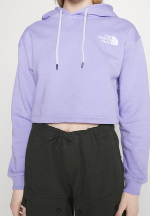 Henkilö pukeutunut lyhennettyyn laventelinväriseen huppariin, jossa valkoiset nyörit ja The North Face -logo, yhdistetty mustiin korkeavyötäröisiin housuihin.