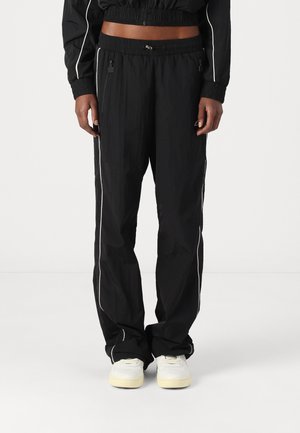 Black track pants with a loose fit, elastic waistband, side zip pockets, and white piping accents down the sides. Made from a lightweight material.