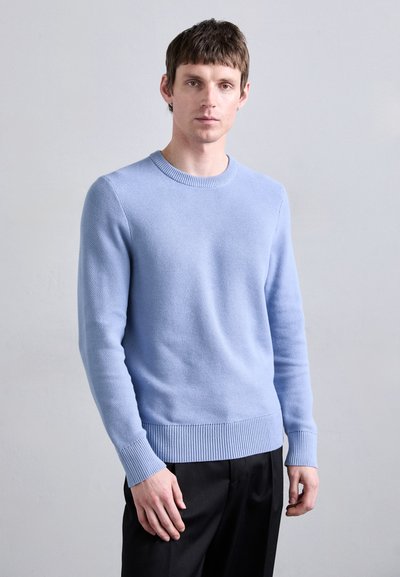 Pull en maille bleu clair avec un col rond et des poignets côtelés. La texture est respirante avec une finition lisse. Associé à un pantalon sombre.