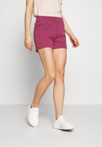 Rebrované pletené šortky v tmavě růžové barvě s širokým elastickým pasem, kombinované s bílými teniskami. Jednoduchý a přiléhavý střih.