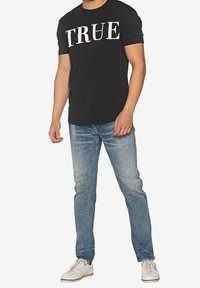 Černé bavlněné tričko s velkým bílým nápisem „TRUE“, doplněné světle modrými roztrhanými džínami a bílými teniskami.