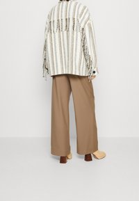 Giacca a frange a righe in bianco, giallo e marrone abbinata a pantaloni palazzo beige e scarpe con tacco block beige. Dettagli del tessuto texture visibili.