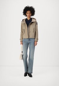 Giacca con cappuccio beige con chiusura a bottoni, due tasche laterali e un patch con logo sulla manica, abbinata a jeans azzurri chiari e stivali neri.
