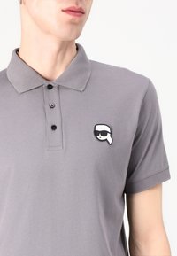 Pelēkā polo krekls, kas izgatavots no kokvilnas, ar klasisku apkakli, trim melnām pogām un mazu krāsainu logo uzlīmi, kurā attēlotas saulesbrilles.