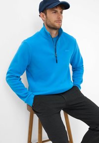 Pullover in pile blu con zip, colletto alto, maniche lunghe e dettagli con logo. Abbinato a pantaloni grigi scuri, seduto su uno sgabello di legno.