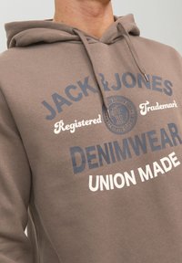 Sudadera marrón hecha de una mezcla de algodón. Presenta texto impreso en azul oscuro y blanco, incluido el nombre de la marca y "DENIMWEAR UNION MADE."