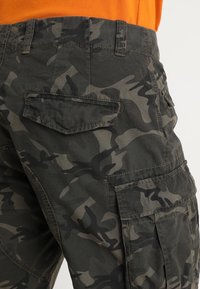 Pantaloni cargo camouflage con un motivo verde scuro e nero, dotati di una tasca posteriore e un tessuto testurizzato; progettati per resistenza e funzionalità.