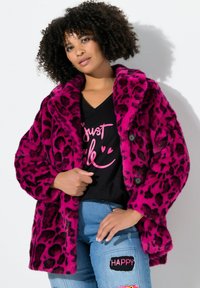 Abrigo de piel sintética fucsia con estampado de leopardo negro, gran cuello y dos botones. Llevado sobre una camiseta negra y unos vaqueros azules con parches.
